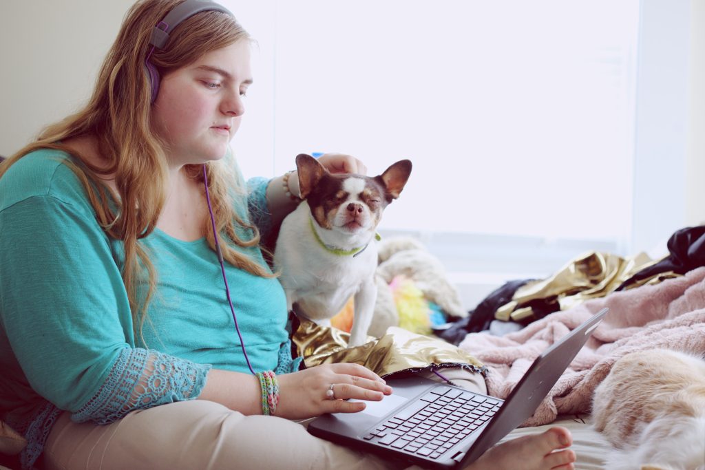 Image of a woman sitting and looking at a laptop balance on her legs. A small dog is next to her and the woman is patting it.