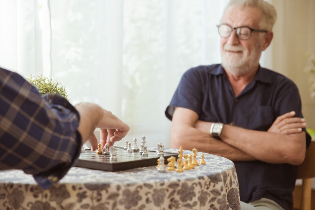 An older man is looking at a chess board with arms crossed as his chess partner, of whom we can only see an arm,makes a move,