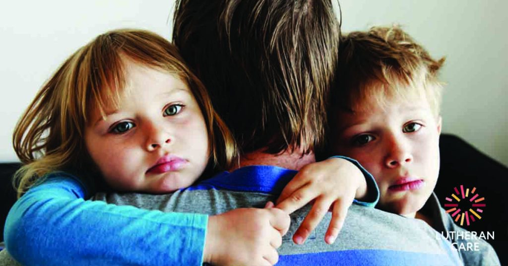 Image of a man’s back he is holding his two young children who are facing the camera in his arms. The Lutheran Care logo appears in the bottom right hand corner.