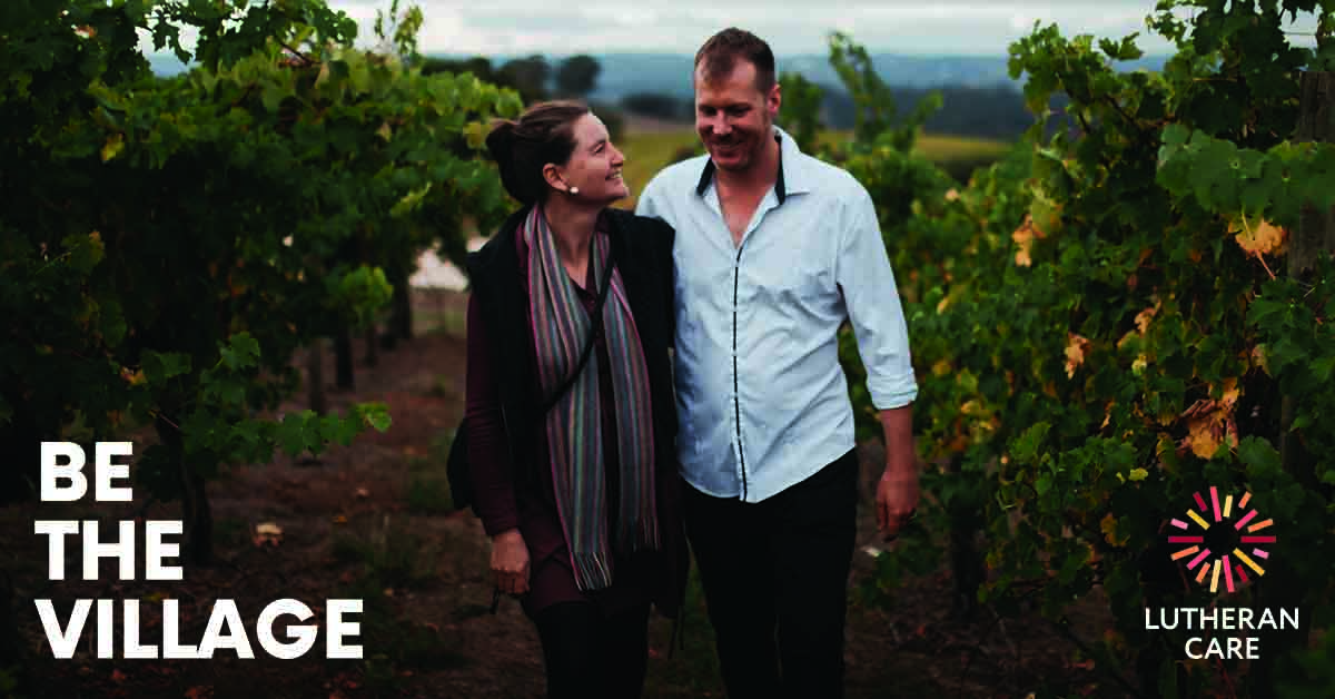 Respite Carers Amanda and Sean walking in a vineyard smiling. The Be The Village and Lutheran Care logos appear at the bottom of the image.
