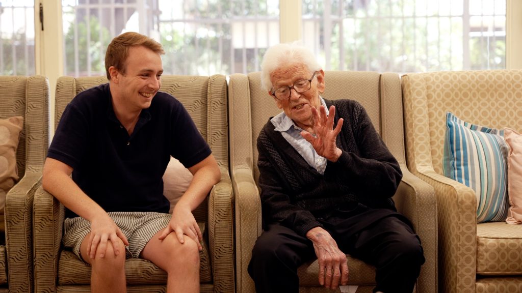 A younger man wearing a dark shirt and light shorts smiles, whilst sitting next to an older man wearing dark pants and a dark jumper over a lighter collared shirt. The older man is speaking animatedly whilst gesturing with his left hand. The two men are part of the Community Visitors Scheme.
