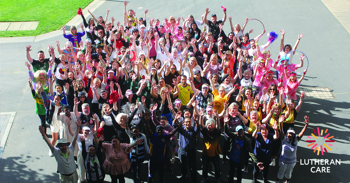 Over 100 Lutheran Care staff members at the 2022 Staff Day standing together outside in costumes smiling with arms raised at camera above the group. The Lutheran Care logo appears in the bottom right hand corner.