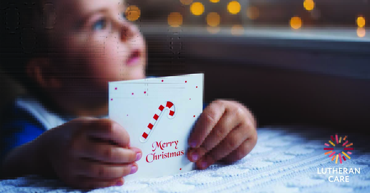 Image of young child holding a card that says Merry Christmas. The Lutheran Care logo appears in the bottom right hand corner.