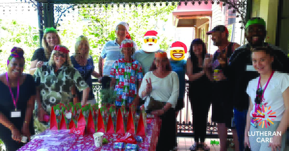 Image of twelve people wearing Christmas party hats gathered around a table with Christmas gifts on it outside. The Lutheran Care logo appears in the bottom right hand corner.