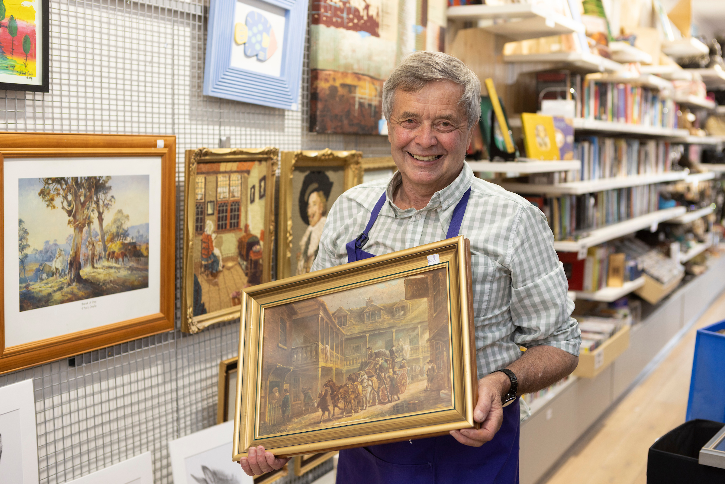 An older man is standing in a Elcies Op Shop holding a painting