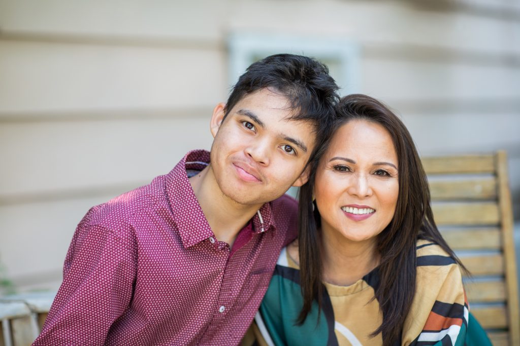 Young adult man sits outside with his mother who is smiling at the camera.