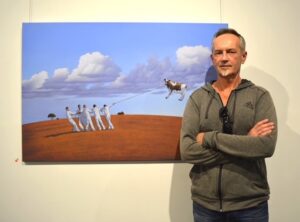 Artist Andrew Baines wears grey hooded jacket. He has his arms crossed and stands in front of one of his surreal paintings, featuring a cow which is flying in the air, tethered by a rope being held by some figures wearing white.