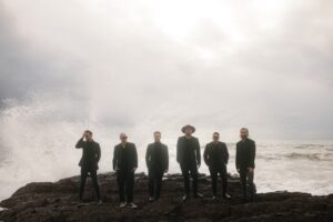 The band Motel Music comprises six men wearing dark clothing standing on a rock in front of a white and stormy ocean. 