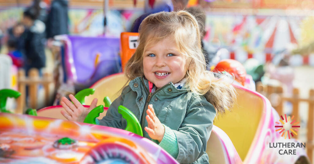 A smiling young girl sits in a show ride. The Lutheran Care logo appears in the bottom right hand corner of the image.