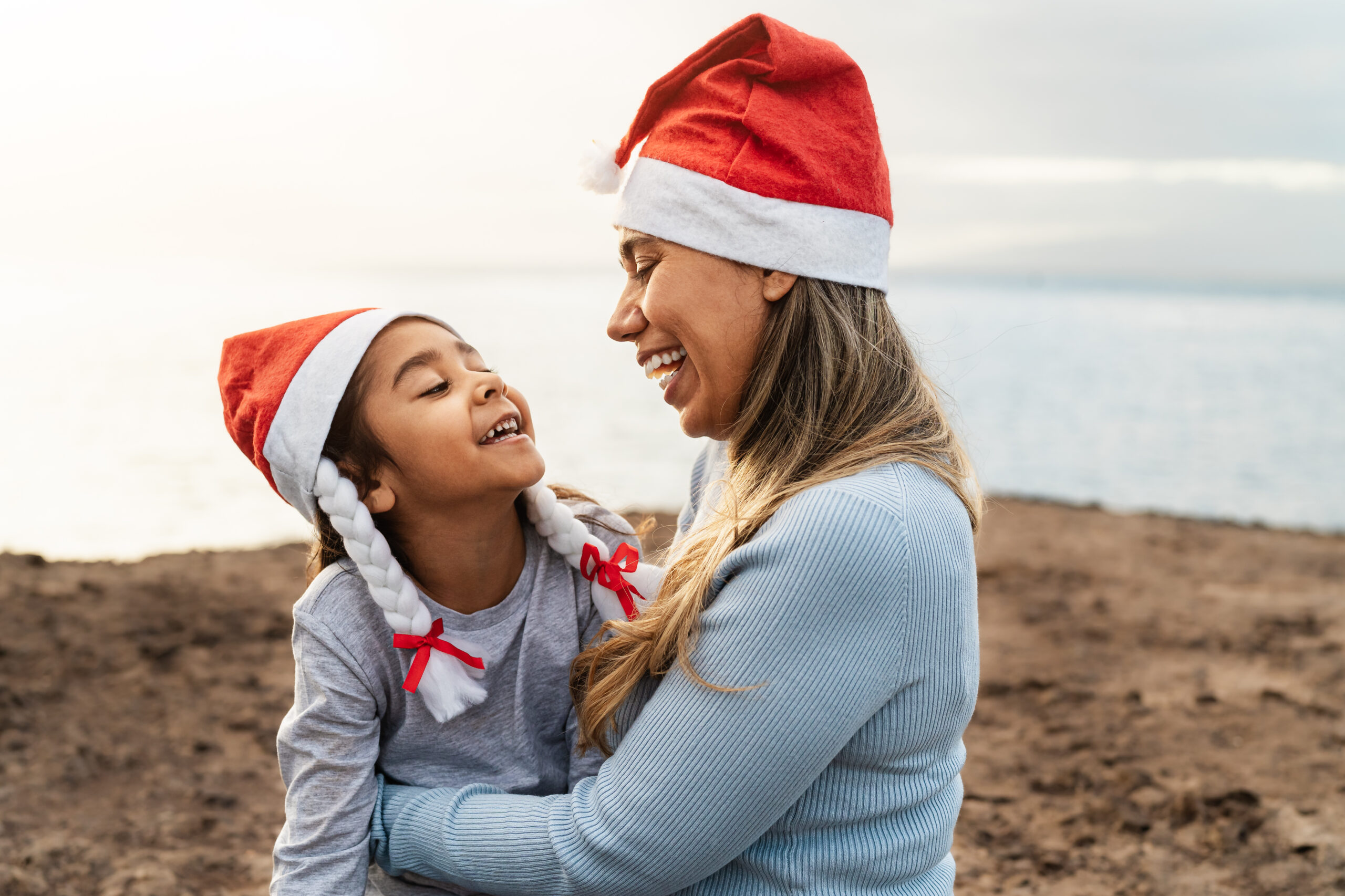 A mother and daughter both wearing Santa hats are smiling at each other near the sea.