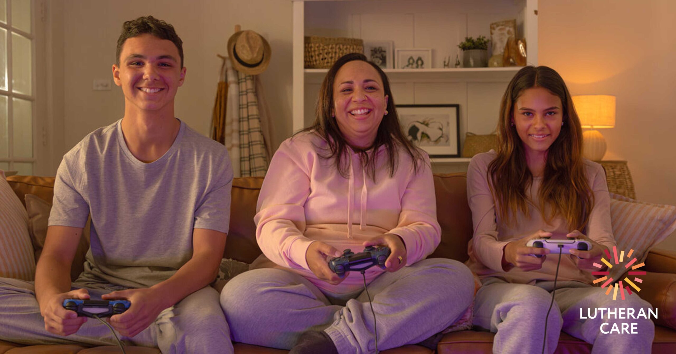Image of a woman and a teenage boy and girl sat on a couch all holding game console controllers. They are smiling whilst playing a game. The Lutheran Care logo appears in the bottom right hand corner.