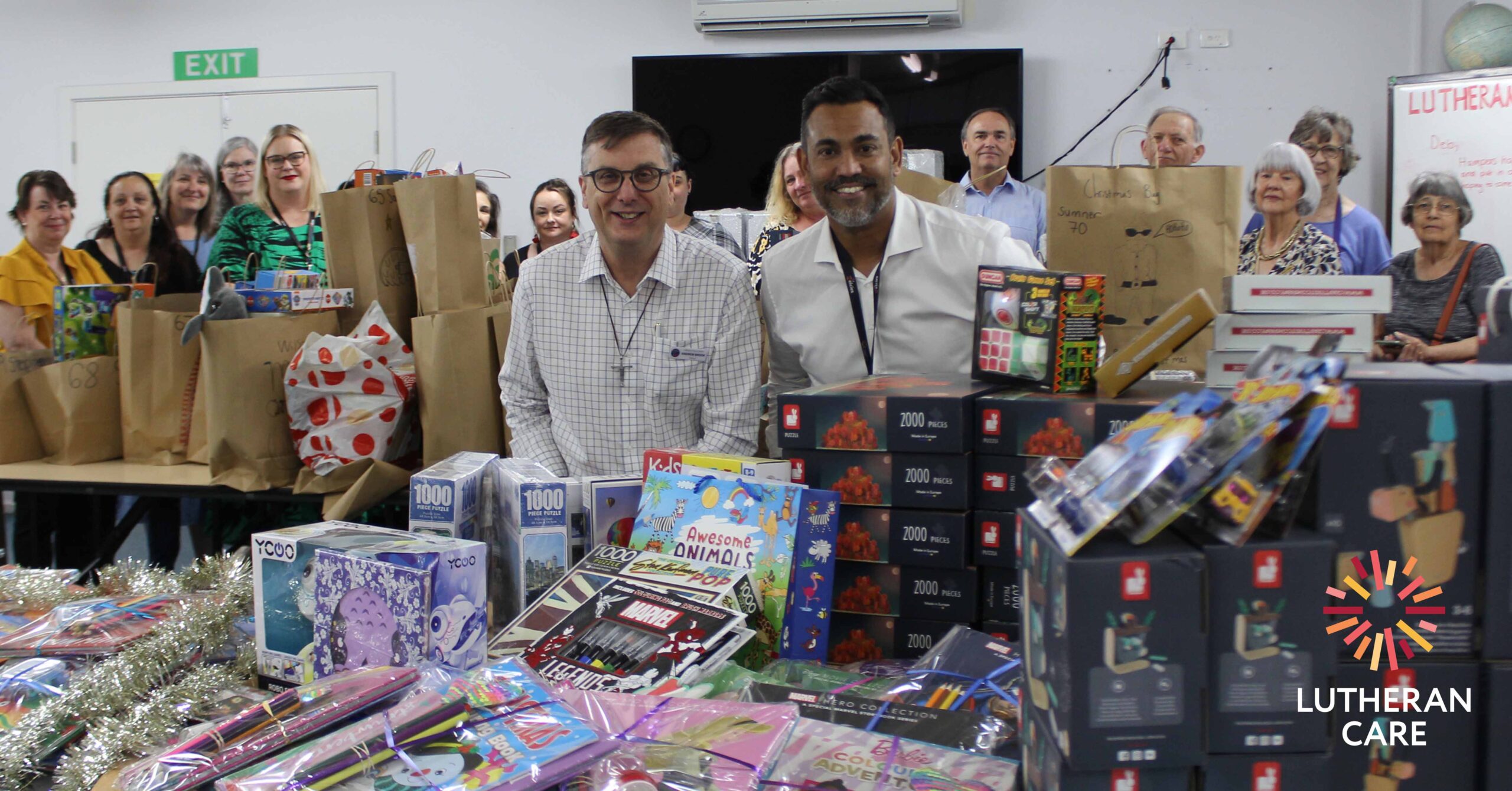 Lutheran Care CEO Rohan Feegrade and Bishop Andrew Brook with staff and volunteers as Christmas Hampers are put together at the Blair Athol offices. The Lutheran Care logo appears in the bottom right hand corner.