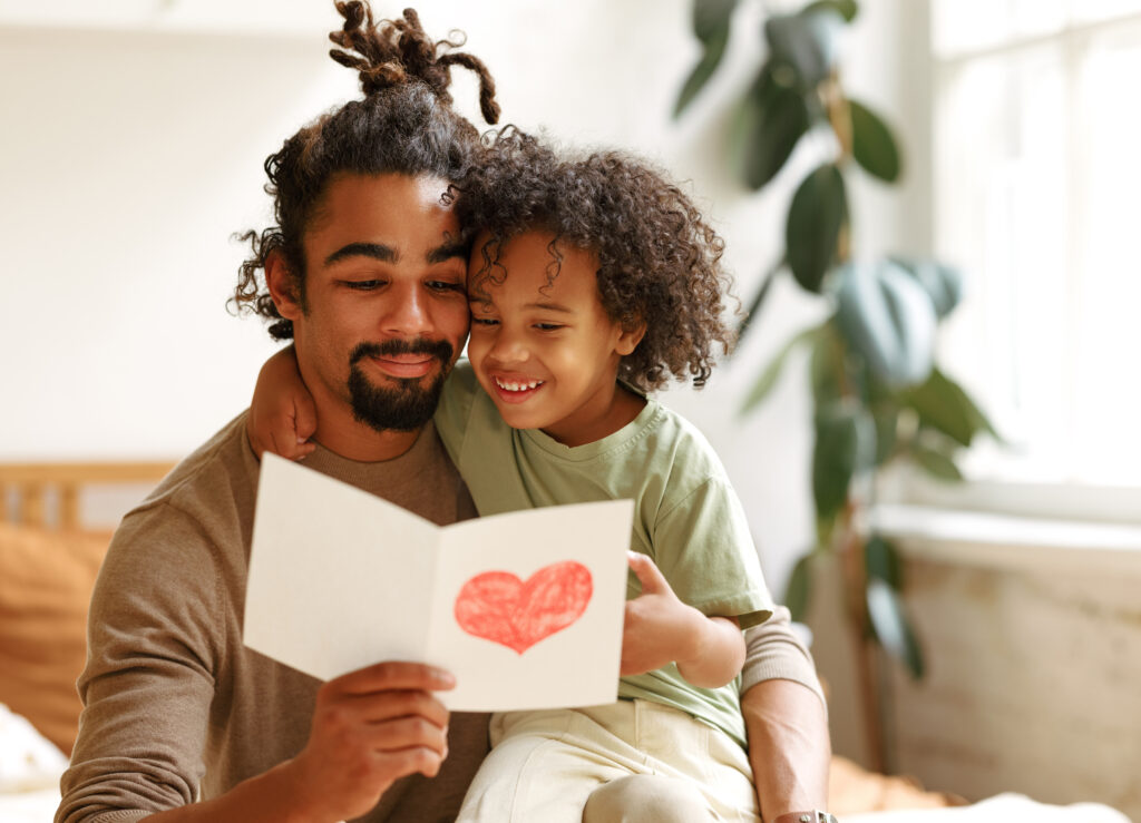 A young boy is sitting with his arm around his father reading the birthday card he has made for him.