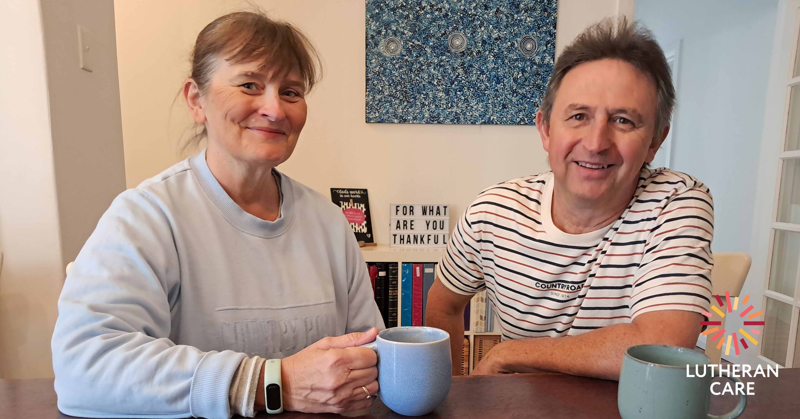 Foster Carers Kristy and Chris sit with a cup of coffee or tea in their home. The Lutheran Care logo appears in the bottom right hand corner.