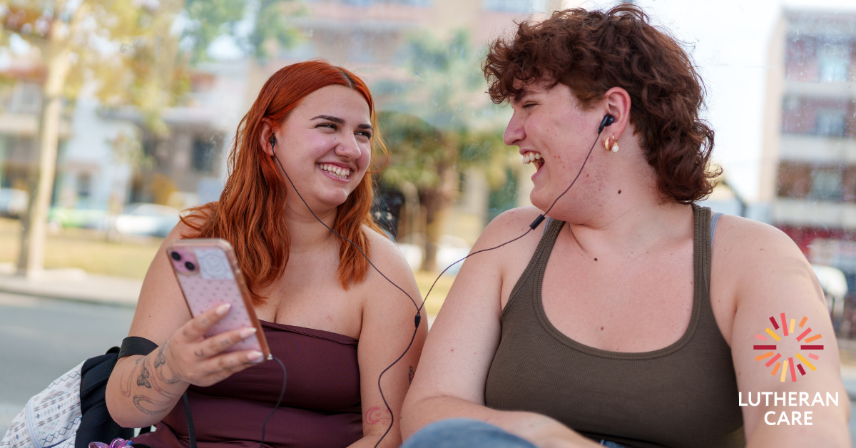 Two young people laugh together outside at something on a mobile phone. The Lutheran Care logo appears in the bottom right hand corner.