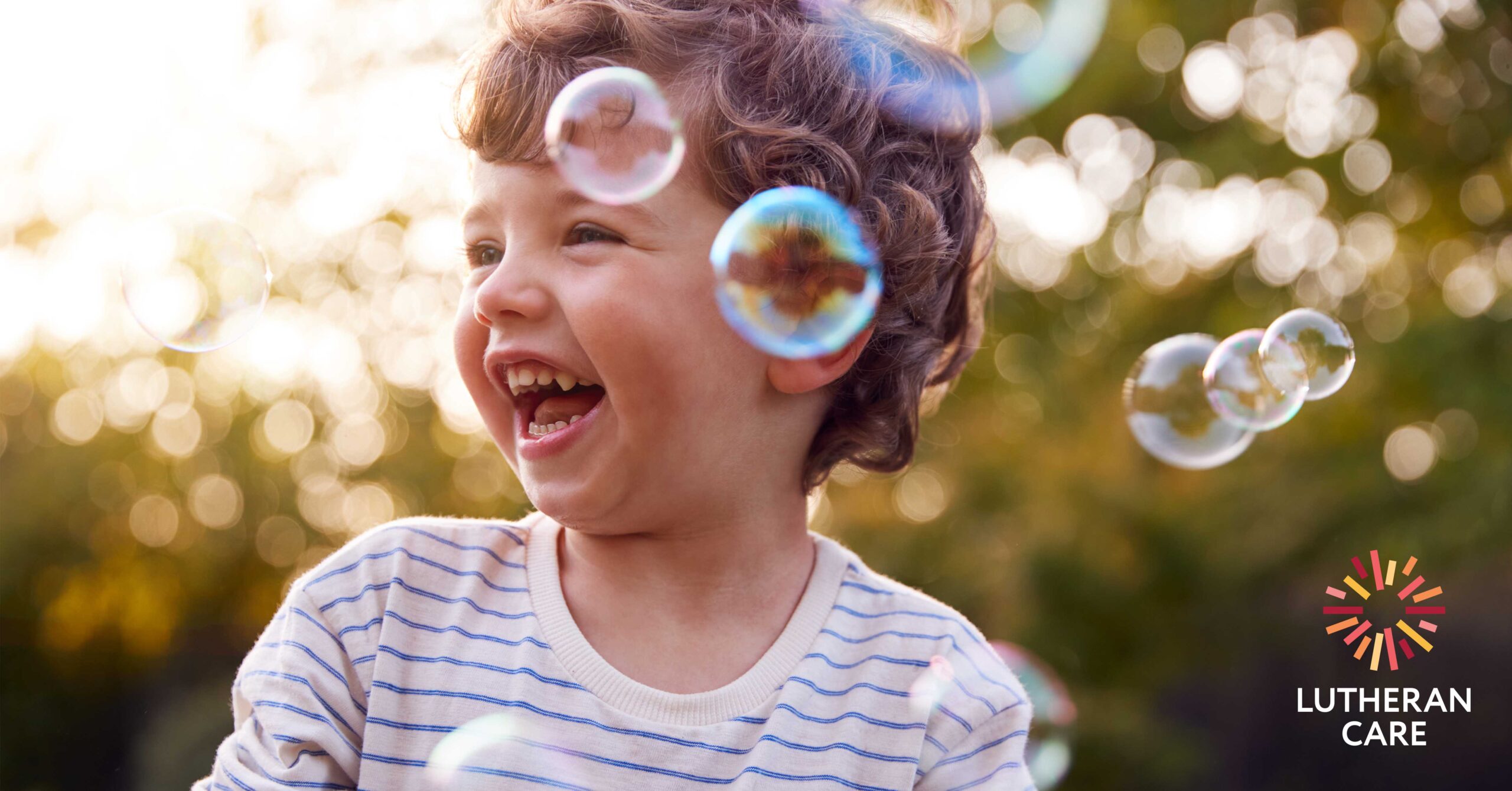 Little boy outside laughs as bubbles float around him. The Lutheran Care logo appears in the bottom right hand corner.