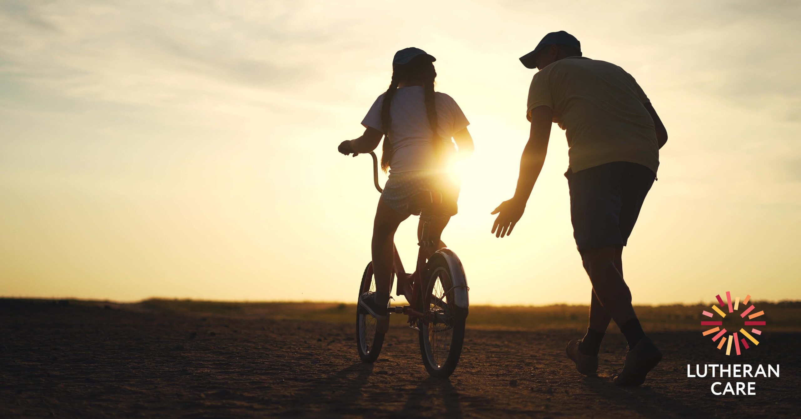 A young girl learns to ride a bike on a dirt road, her guardian runs beside her. The Lutheran Care logo appears in the bottom right hand corner.