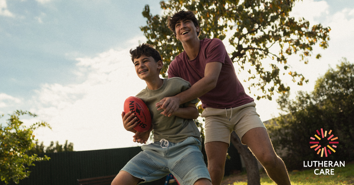 Two teenage boys laughing and playing football in the back garden. The Lutheran Care logo appears in the bottom right hand corner