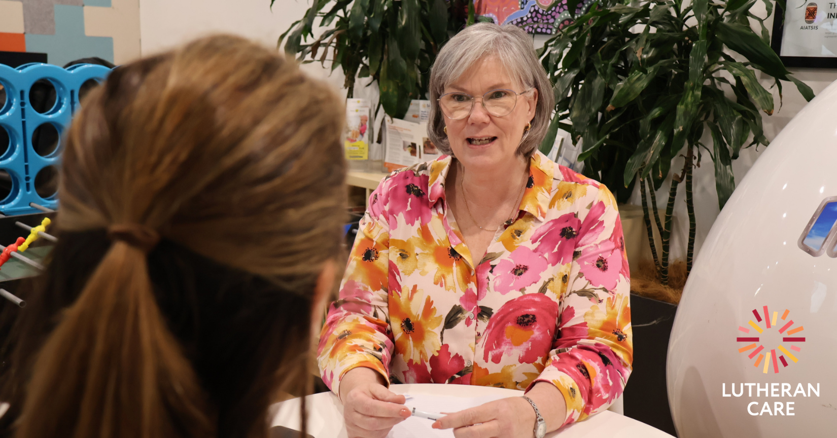 Image of senior financial counsellor Anna speaking with a client who has their back to the camera. The Lutheran Care logo appears in the bottom right hand corner.