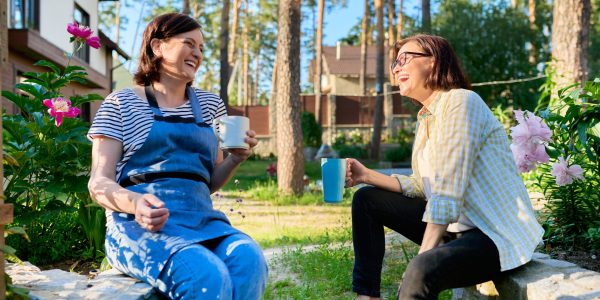 Two women sit outside laughing together drinking cups of tea or coffee.