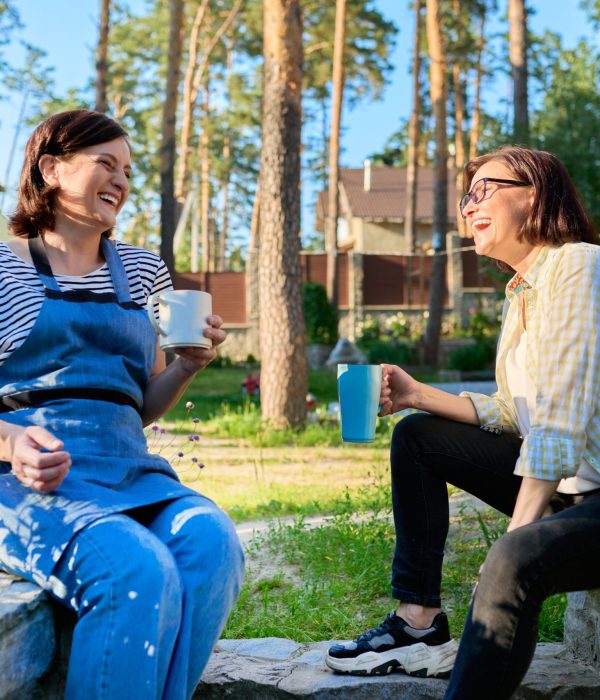 Two women sit outside laughing together drinking cups of tea or coffee.