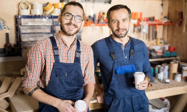 Waist up portrait of two smiling men in a workshop looking at camera while taking break.