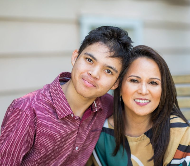 Young adult man sits outside with his mother who is smiling at the camera.