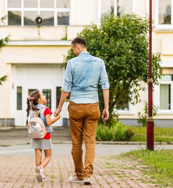 A man and his young daughter are holding hands and talking to each other as they walk towards a building.