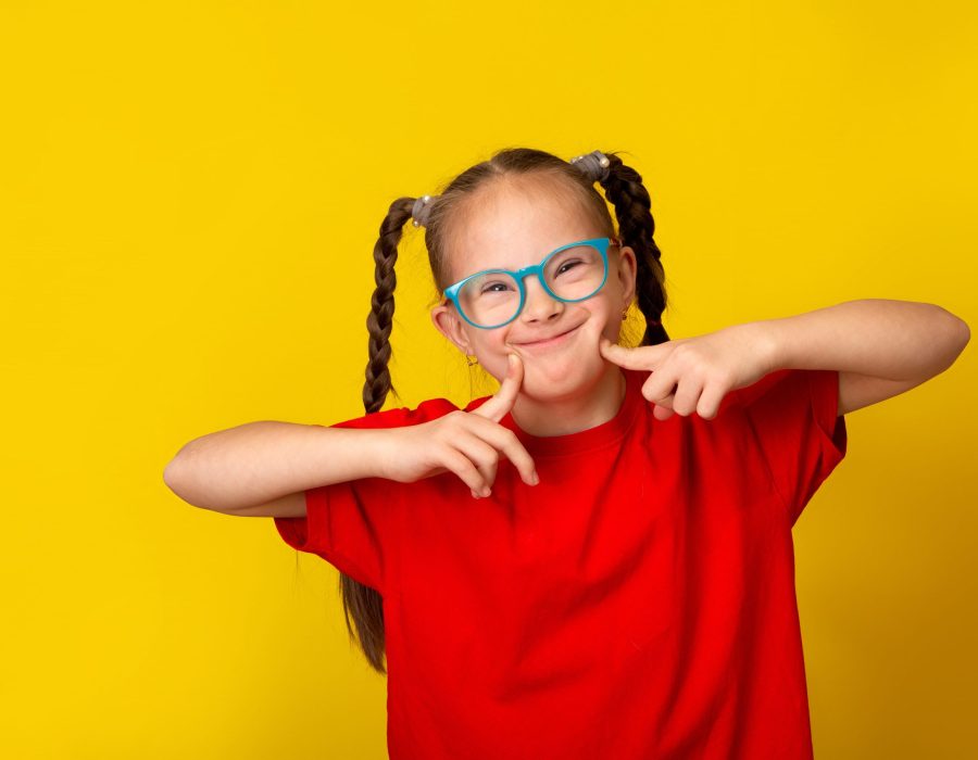 A young girl with Down Syndrome is smiling and has her arms raised and a finger from each hand pressed into each corner of her mouth
