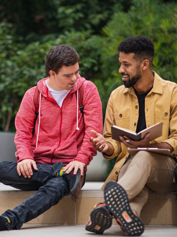 Two young men are sitting on a step outside talking, one man is showing the other something in a book.