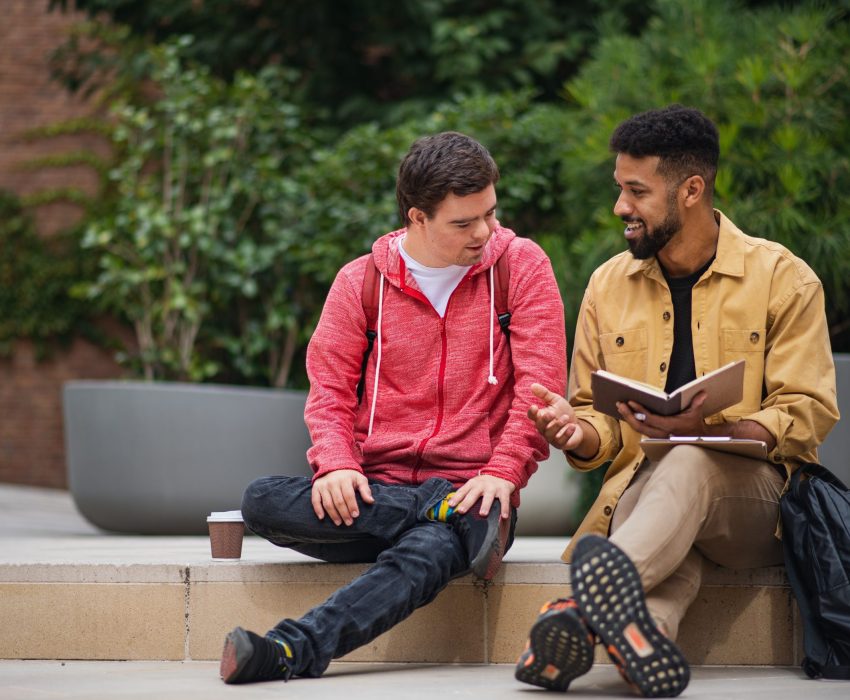 Two young men are sitting on a step outside talking, one man is showing the other something in a book.