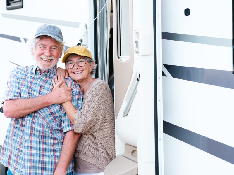 Cheerful senior couple on leisure trip standing outside a camper hugging each other and looking at camera. Beautiful Caucasian couple of retirees enjoying freedom and alternative life in motor home