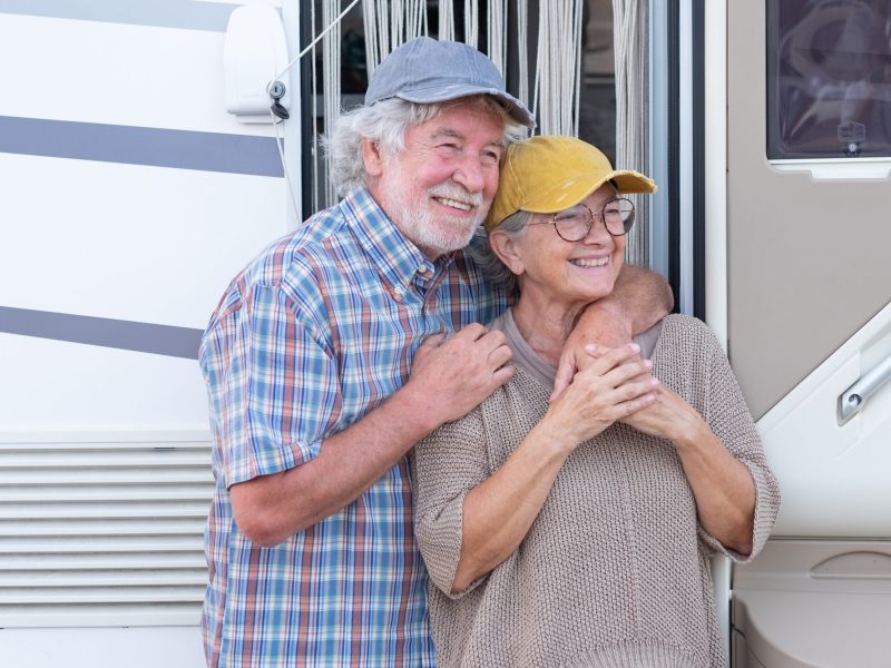 Cheerful senior couple on leisure trip standing outside a camper hugging each other and looking away. Beautiful Caucasian couple of retirees enjoying freedom and alternative life in motor home