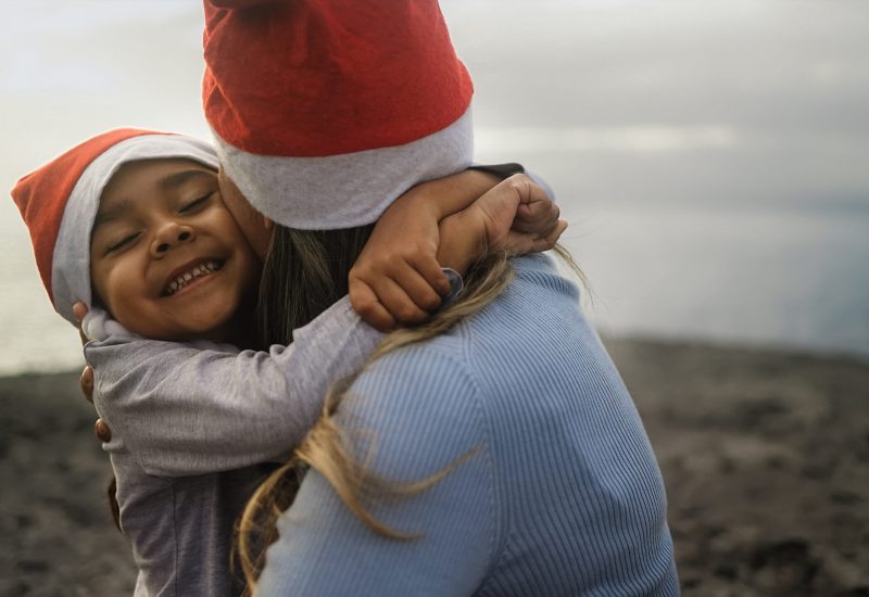 A young girl hugs her mother, both are wearing Santa hats.