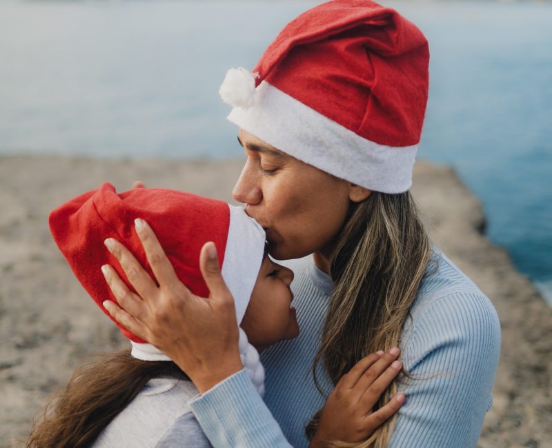 A mother kisses her daughters head, both are wearing Santa hats.