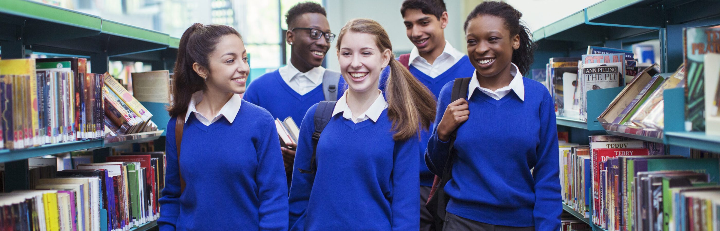 Smiling students wearing blue school uniforms walking between bookshelves in library